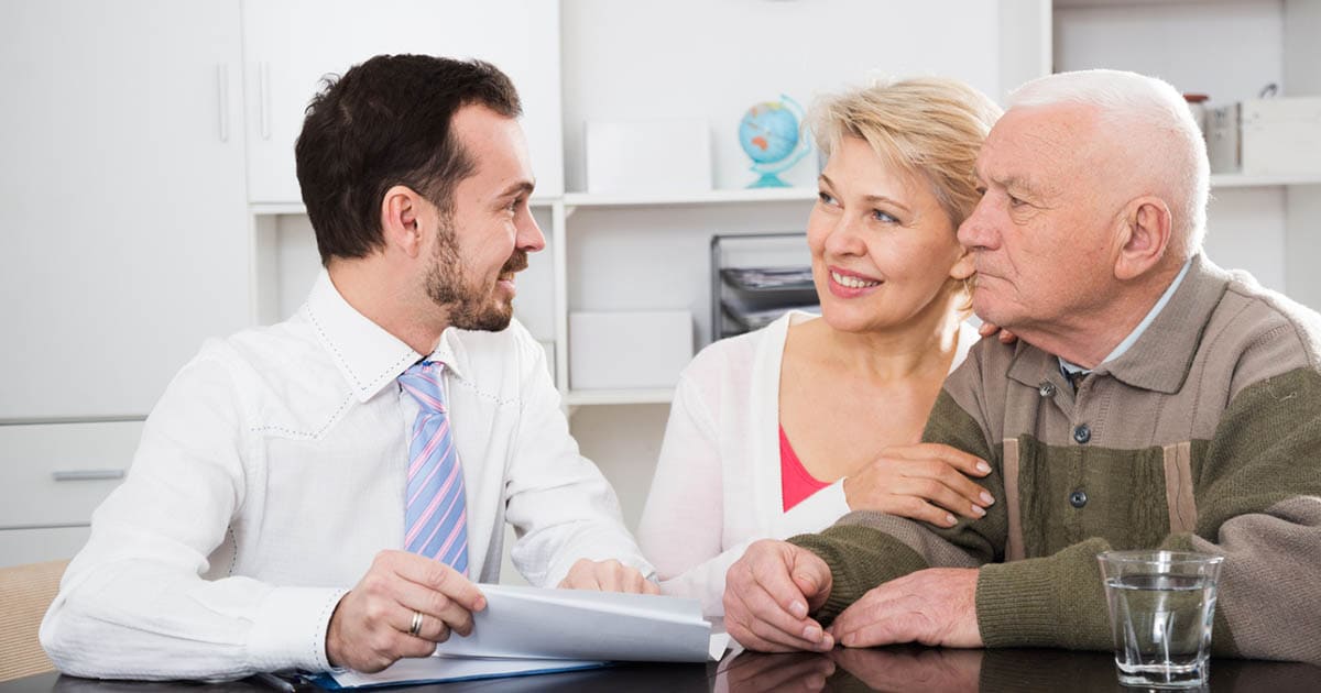 A man in a white shirt and pink tie discusses paperwork with a smiling woman and an older man seated at a desk in an office environment.
