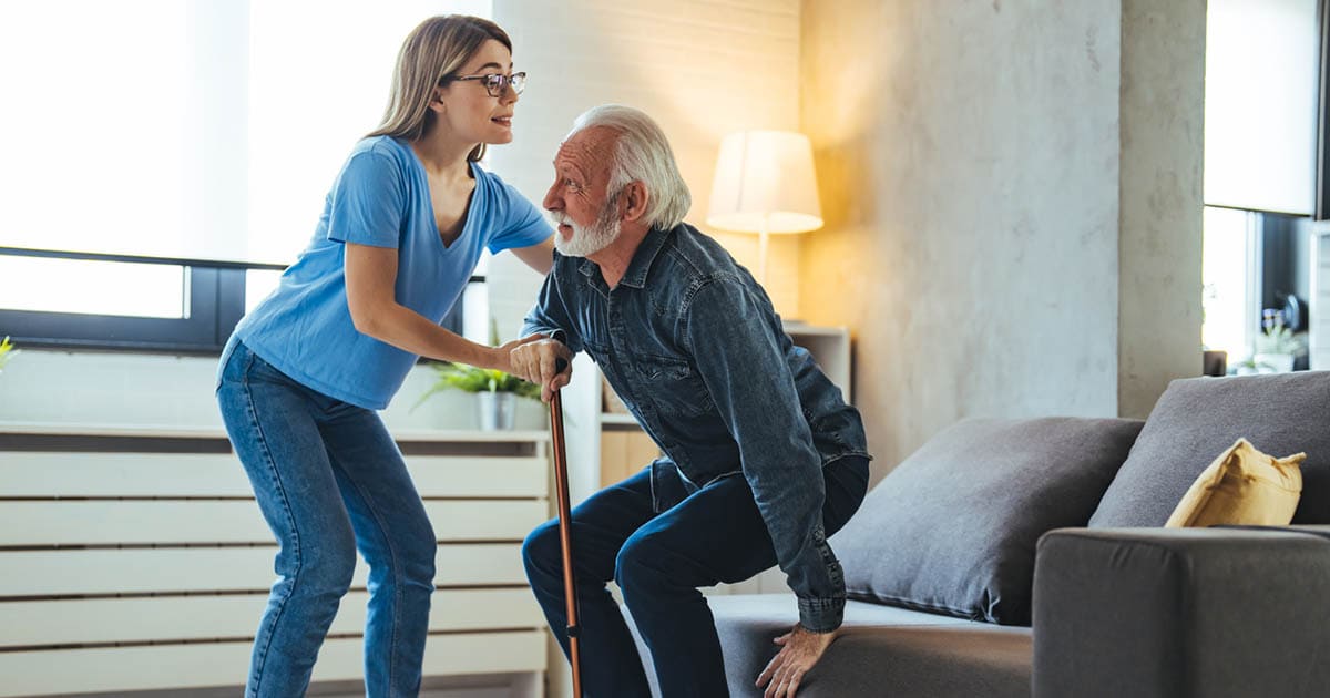 A young woman in a blue shirt assists an elderly man with a cane as he stands up from a couch in a living room. A young woman in a blue shirt assists an elderly man with a cane as he stands up from a couch in a living room.