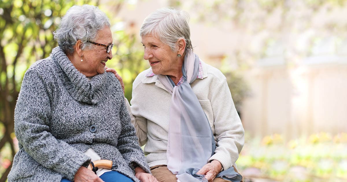 Two elderly women sit outdoors on a bench, smiling and conversing with each other in a garden setting. One wears glasses and a grey sweater, and the other wears a scarf and a light jacket. Two elderly women sit outdoors on a bench, smiling and conversing with each other in a garden setting. One wears glasses and a grey sweater, and the other wears a scarf and a light jacket.