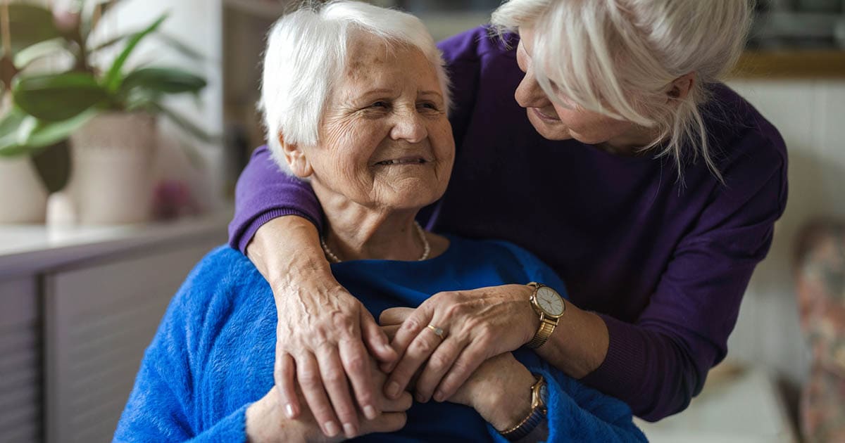 A senior woman sitting, wearing a blue top, is embraced by another woman in a purple top, smiling affectionately at her. Plants are visible in the background.