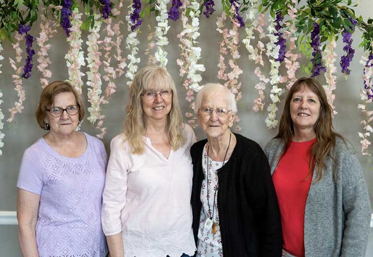 Four women, who reside in senior living in Arcola, IL, stand in front of a floral backdrop. One wears a purple top, another a white blouse, the third dons a black cardigan, and the fourth sports a red shirt with a gray sweater.