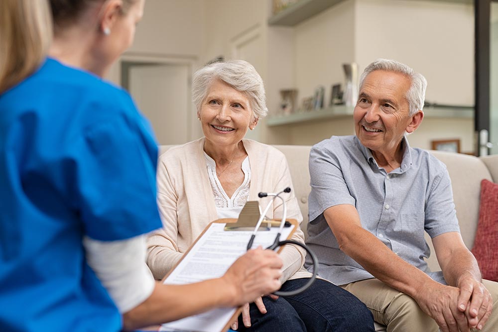 An elderly couple sits on a couch, smiling and talking with a healthcare professional in blue scrubs who holds a clipboard and stethoscope, discussing senior living in Illinois. An elderly couple sits on a couch, smiling and talking with a healthcare professional in blue scrubs who holds a clipboard and stethoscope, discussing senior living in Illinois.